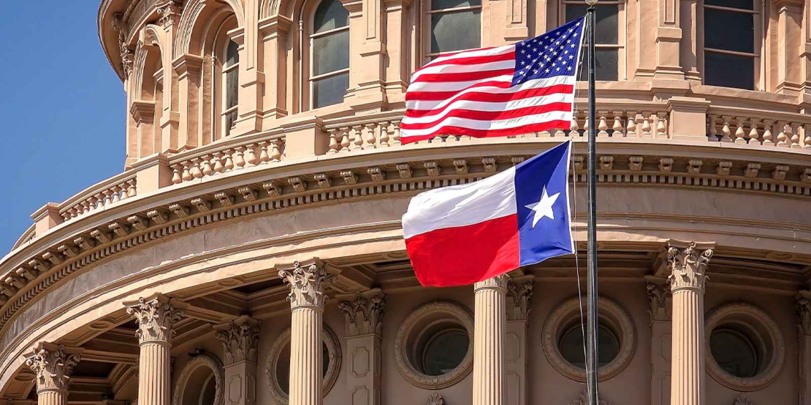 American and Texas state flags flying on the dome of the Texas State Capitol building.