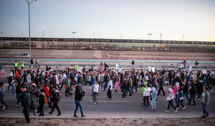 Photo: A large crowd streams by a a freeway and border fencing. They carry signs and american flags, walking in a general procession. 
