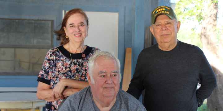 Image: A group of three pose for a photo outside of a wooden house, two standing and one sitting. The woman leans on the chair one man is sitting on. The man standing wears a baseball cap indicating he is a Vietnam War vet. 
