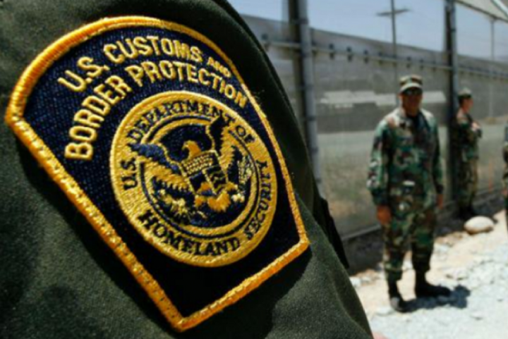 Photo: A Customs and Border Protection patch is worn on a green uniform. The CBP officer's arm takes us most of the photo, a fence and another CBP officer stand in the background.