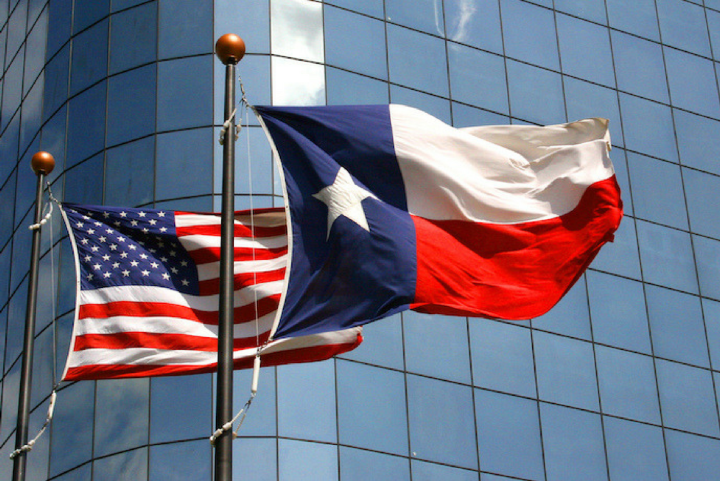 Photo: A Texas and United States flag wave in the wind on two separate flagpoles in front of a corporate building.