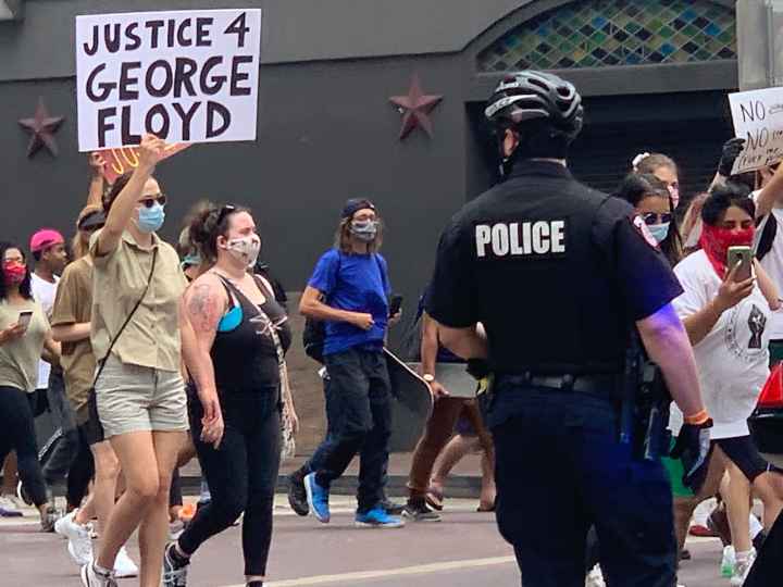 Photo: A police officer looks on as a crowd of people march past at a downtown Houston intersection. In focus is a person holding up a sign that says 'Justice 4 George Floyd.'
