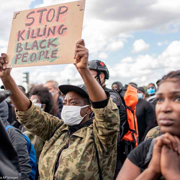 Photo: A crowd of people is gathered and faces one direction. The camera focuses on one African-American person wearing a mask and holding up a sign with handwritten letters that says, "Stop Killing Black People."