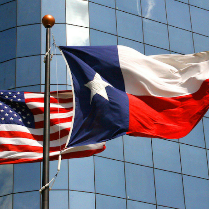Photo: A Texas and United States flag wave in the wind on two separate flagpoles in front of a corporate building.