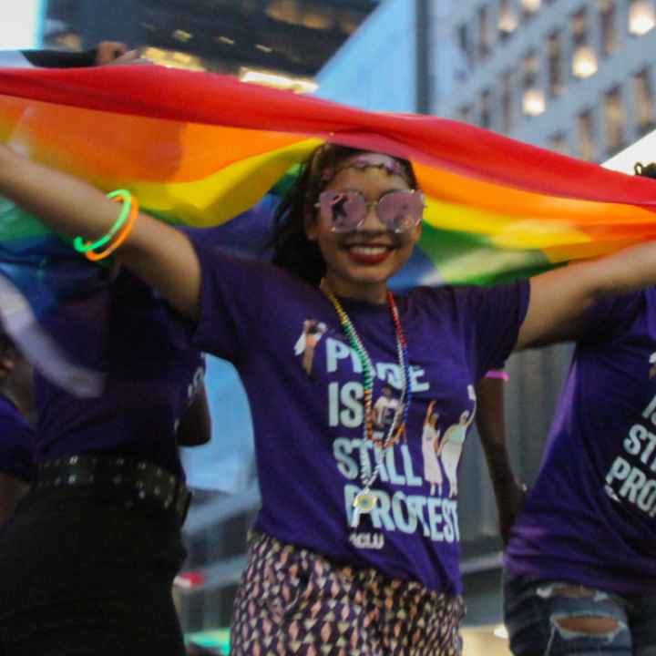 Smiling woman standing with rainbow flag above her head wearing t-shirt that says Pride is Still Protest